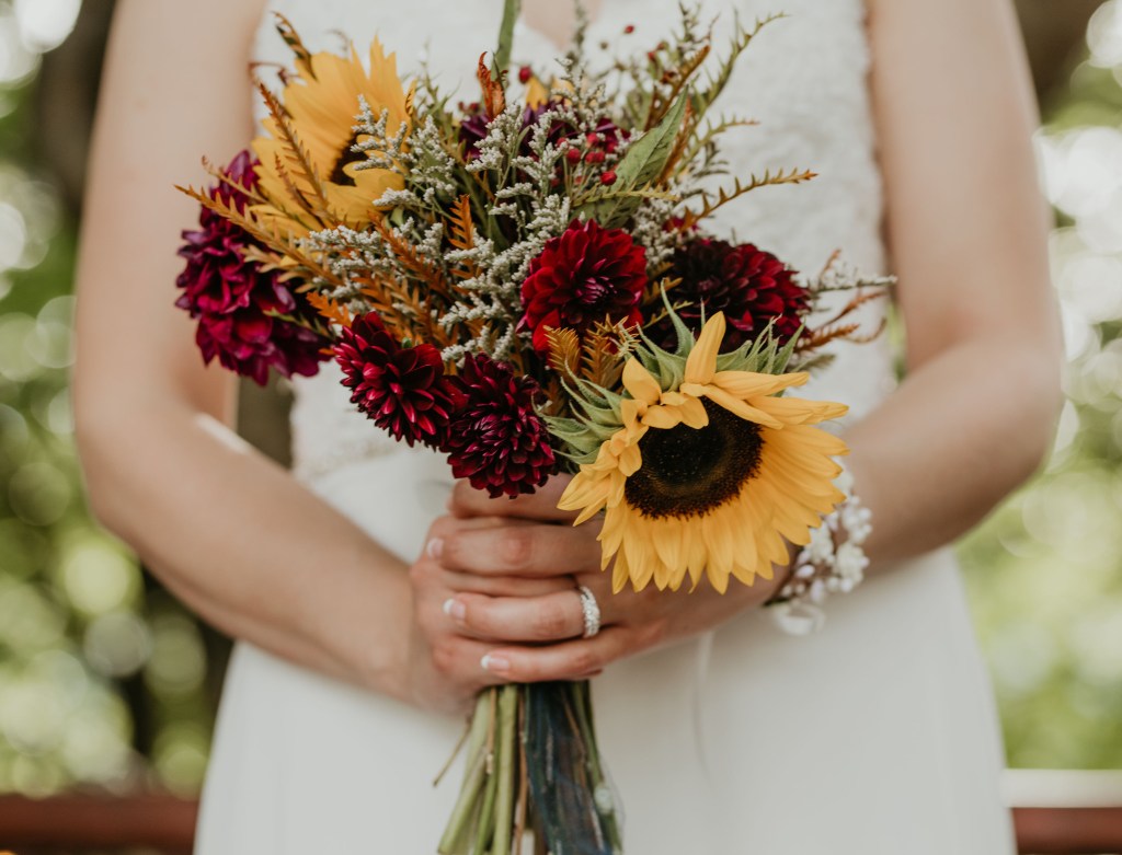 A close-up of a bride holding a vibrant bouquet featuring sunflowers, deep red dahlias, and golden foliage, set against a blurred green background.