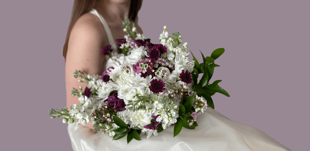 A close-up of a woman holding a large bouquet of white and purple flowers, against a muted purple background.