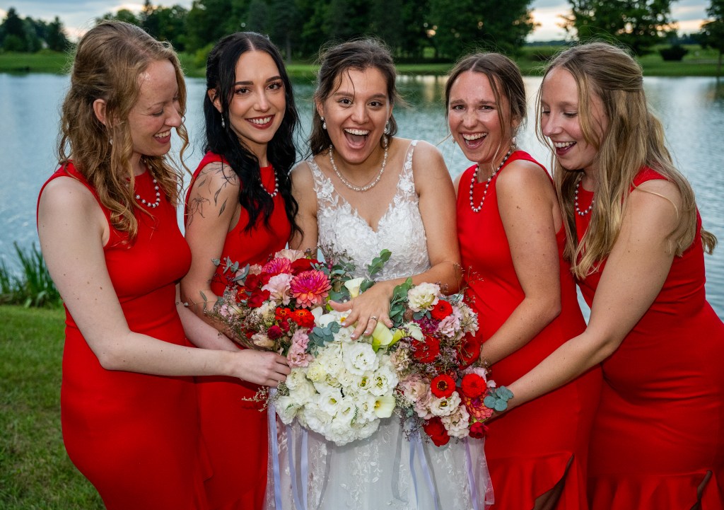 A joyful bride surrounded by her four bridesmaids, all wearing red dresses, as they celebrate by a serene lake. The bride holds a vibrant bouquet of flowers, and they are all smiling.