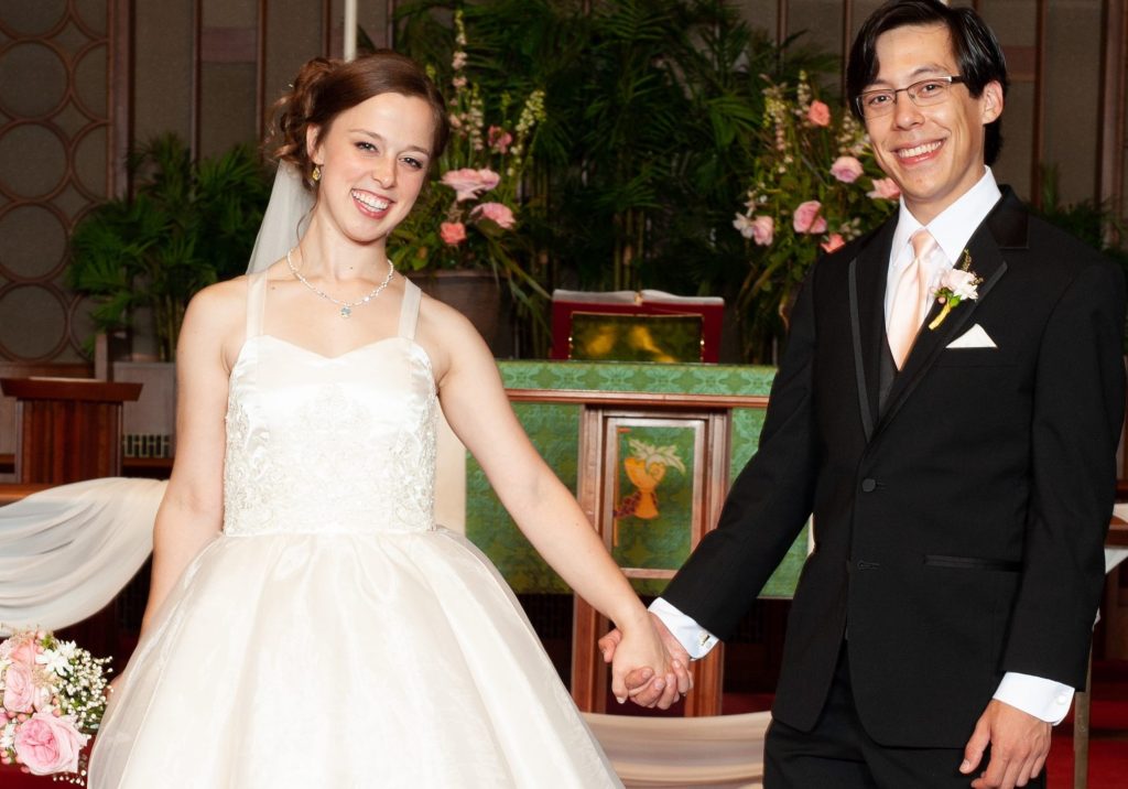 A smiling bride and groom holding hands in a church setting, surrounded by floral decorations.