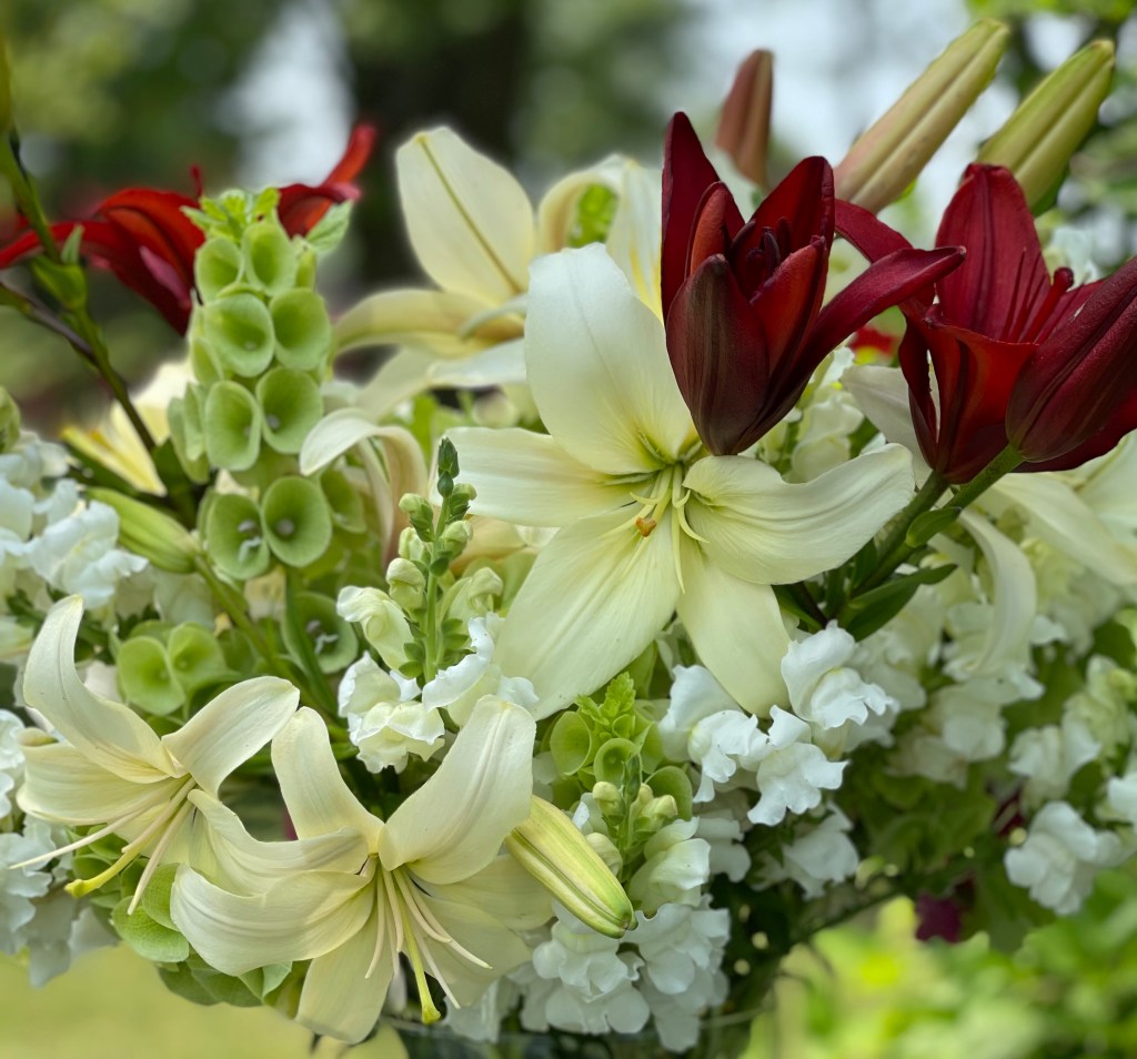 A close-up view of a vibrant bouquet featuring white lilies, red lilies, and various green foliage accents, surrounded by clusters of white flowers.