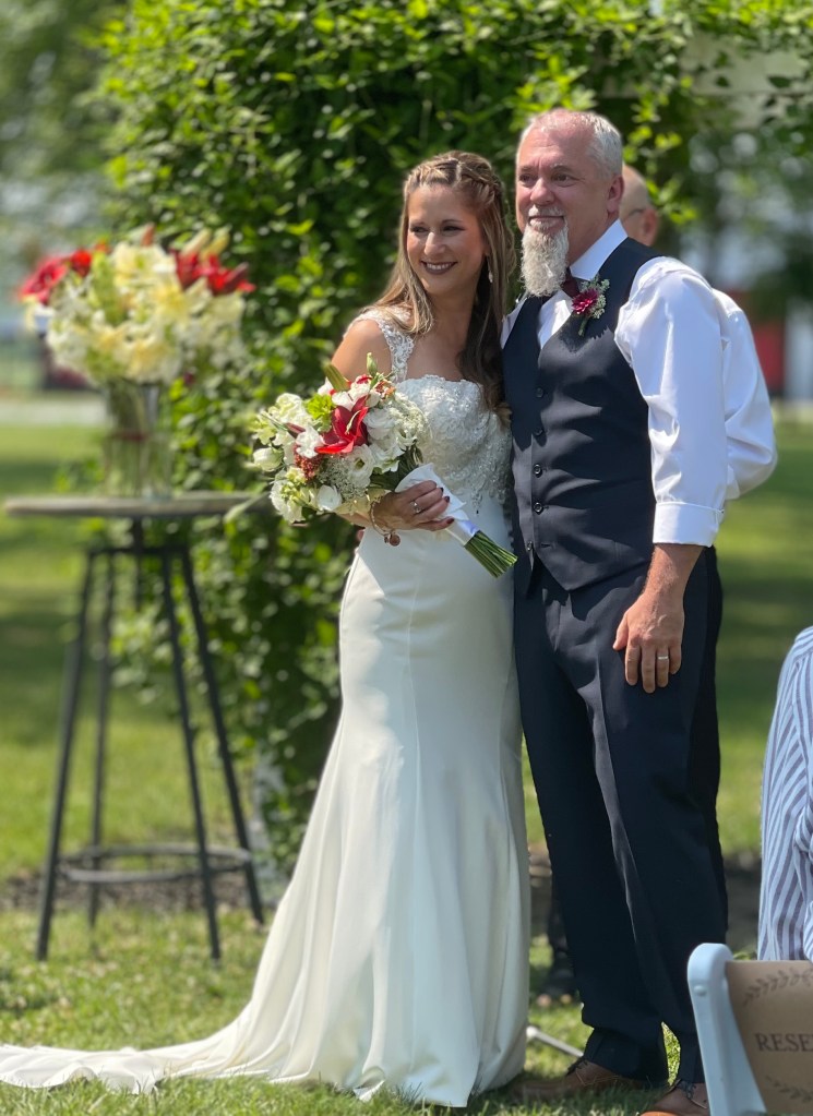 A bride in a white wedding dress stands next to a man in a navy suit, both smiling. The bride holds a bouquet with red and white flowers, with a greenery backdrop.