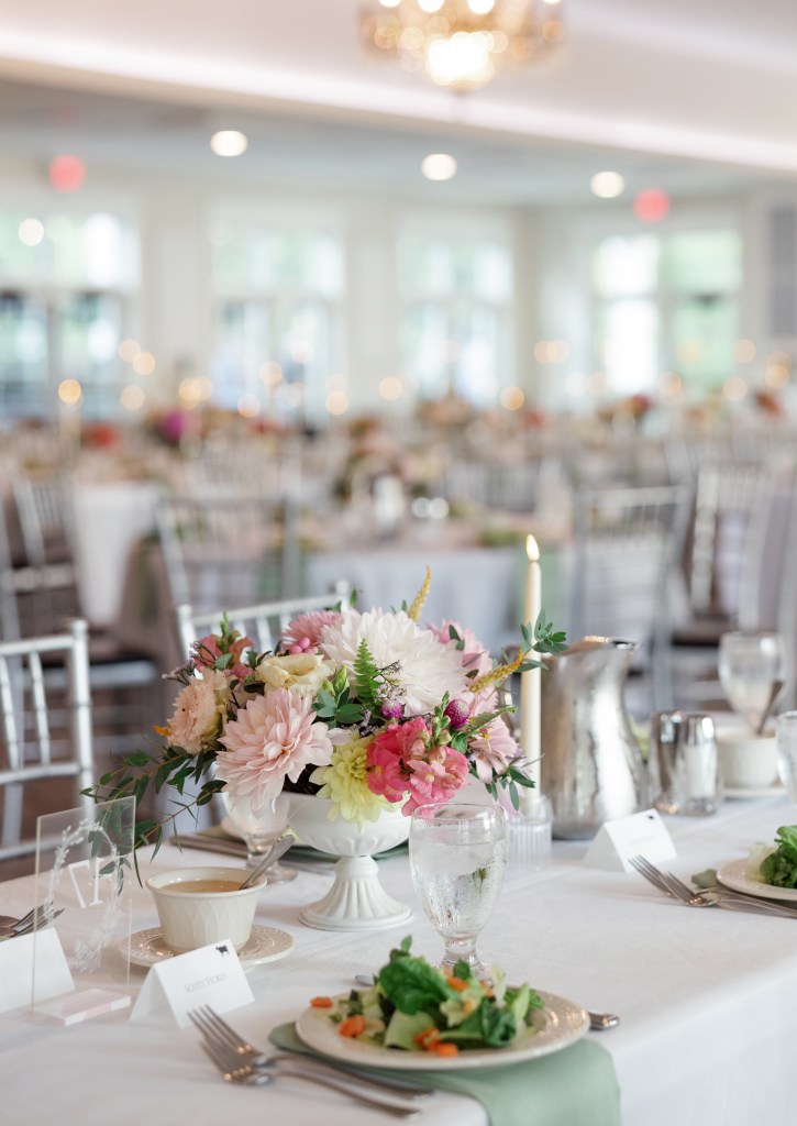 A beautifully set dining table adorned with a large floral arrangement, elegant glassware, and plates featuring salads and soups, in a bright banquet hall.