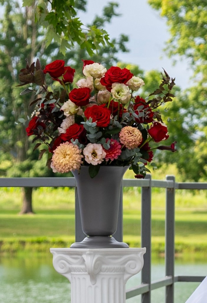 A decorative vase filled with a variety of flowers, including red roses, white blooms, and peach-colored dahlias, positioned on a white pedestal near a serene body of water and surrounded by greenery.